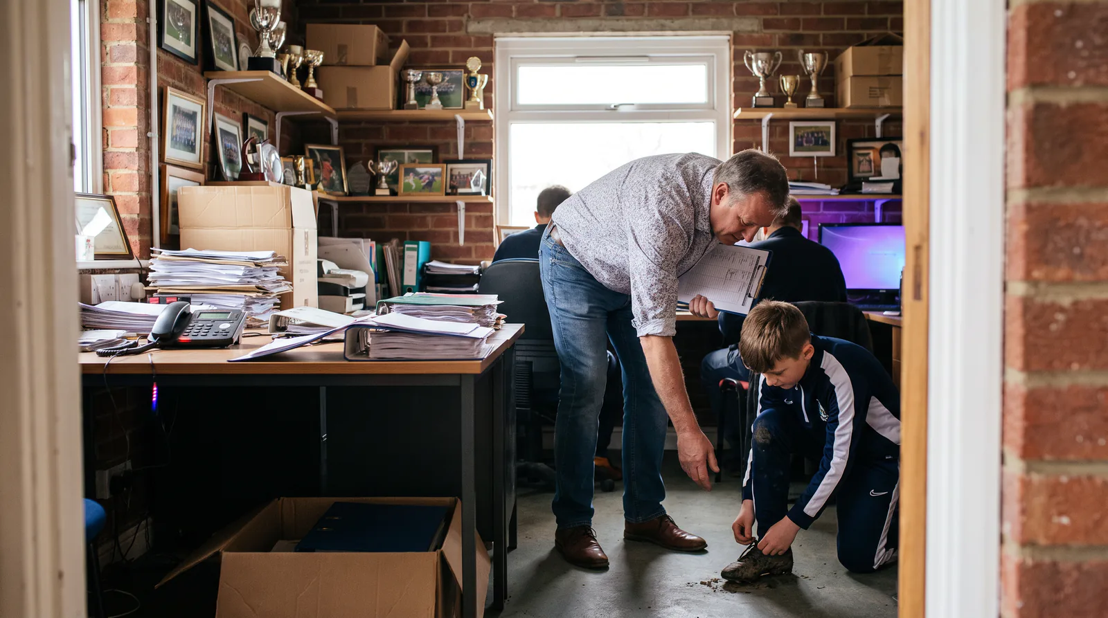 A volunteer in a trophy-lined club office turns away from a ringing desk phone to help a child with their boots